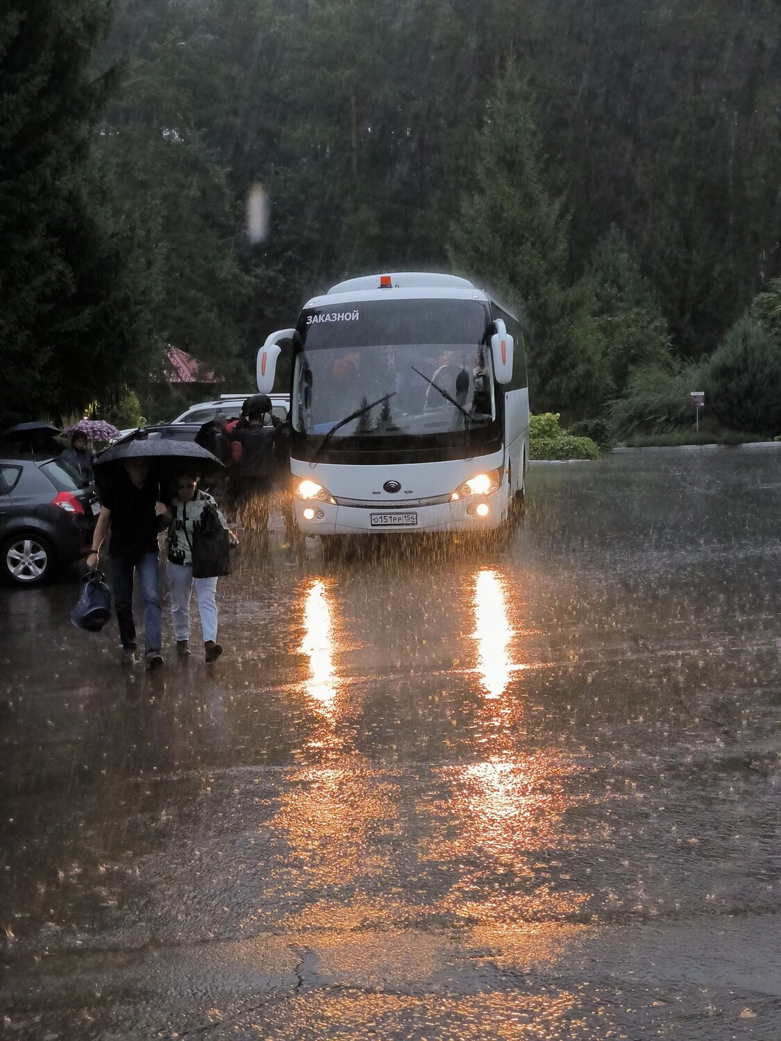 Сцена на улице во время сильного проливного дождя, главным объектом которой является большой белый туристический автобус