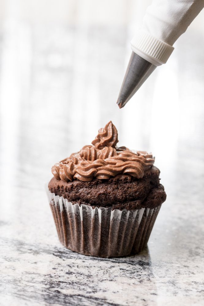 Piping bag with chocolate ganache cream above chocolate cup cake on the grey granite background.