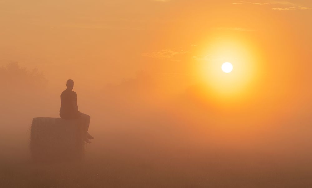 Silhouette of a man sitting on a roll of hay and looking at the sunrise.