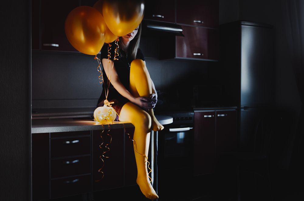 Girl in colored stockings sitting on kitchen table with lantern and balloons in Halloween
