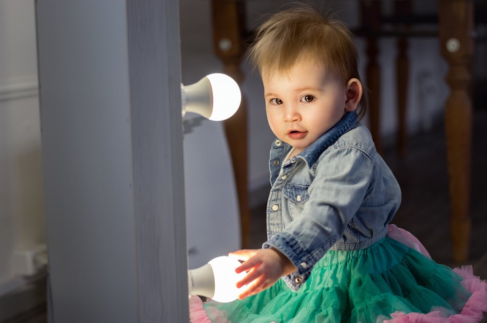 Маленькая знакомая девочка фотографируется у зеркала и держится рукой за светодиодную лампу