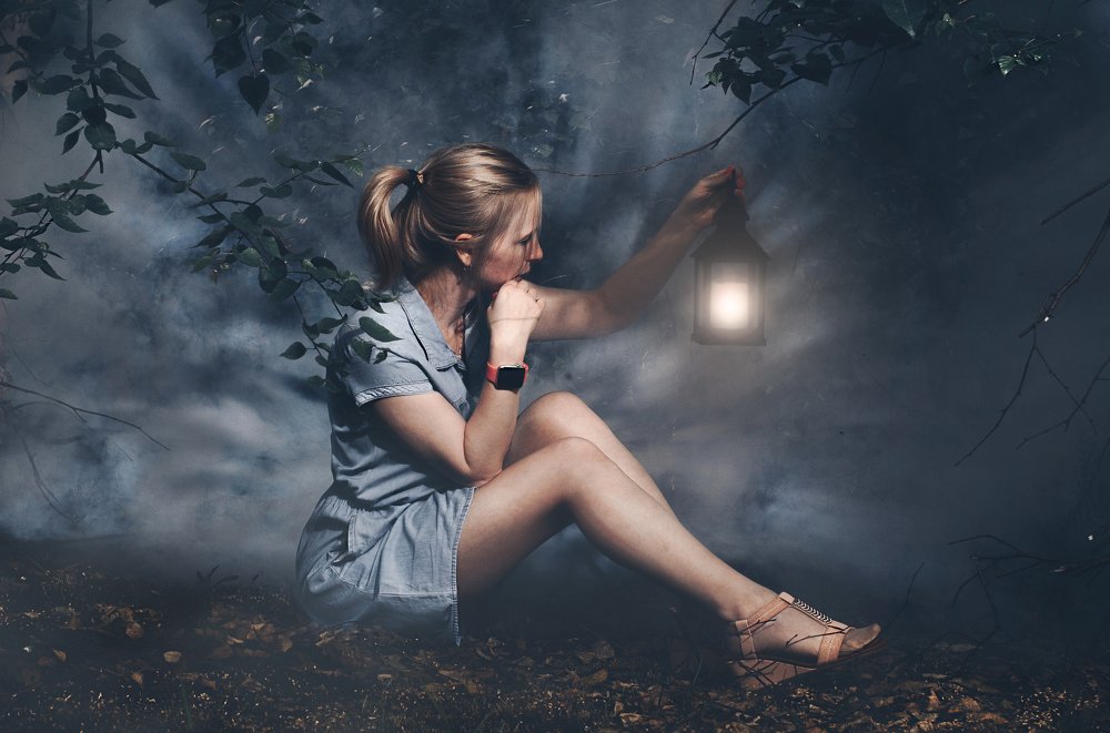 Beautiful girl in denim dress on sitting in forest with fog with lantern in hand