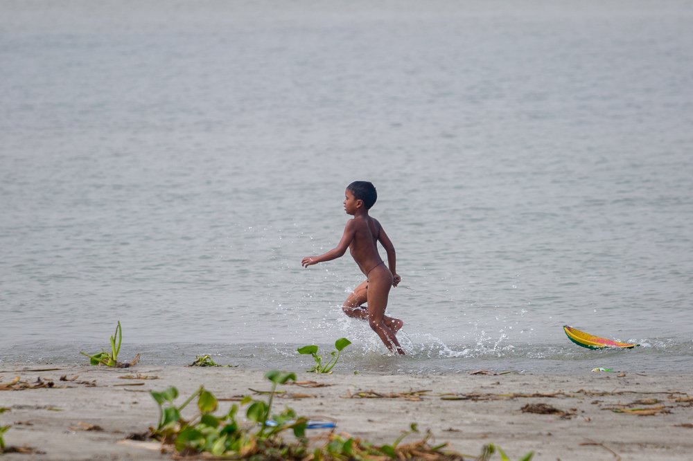 A nude little boy playing with rubber boat in water.